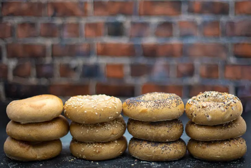 four different kinds of sourdough bagels in stacks in front of a brick wall