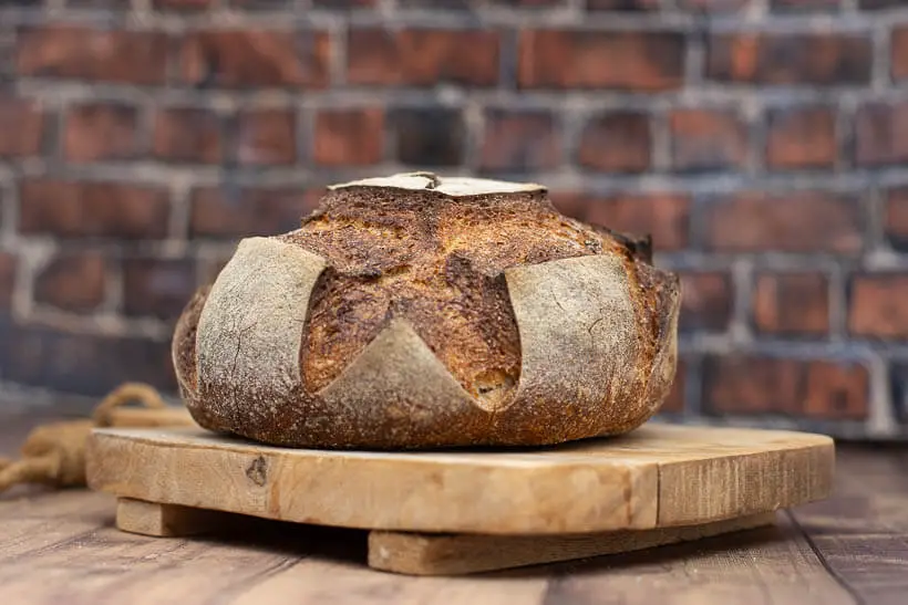 An artisan sourdough boule on a wooden board in front of a brick wall