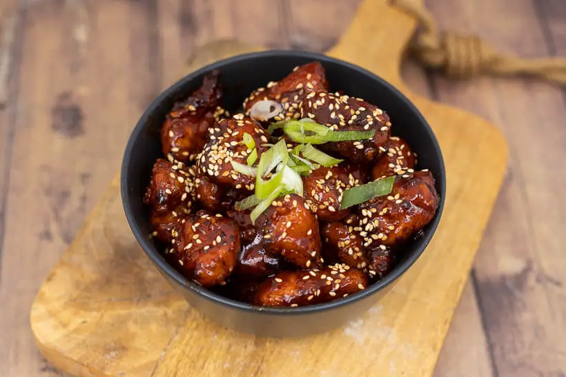 korean fried chicken in a bowl topped with toasted sesame seeds and scallions
